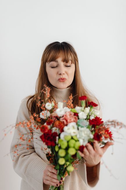 A young woman with light brown hair and bangs, dressed in a beige sweater, is holding a vibrant bouquet of fresh flowers. The bouquet includes red and white roses, blush pink carnations, white ranuncu
