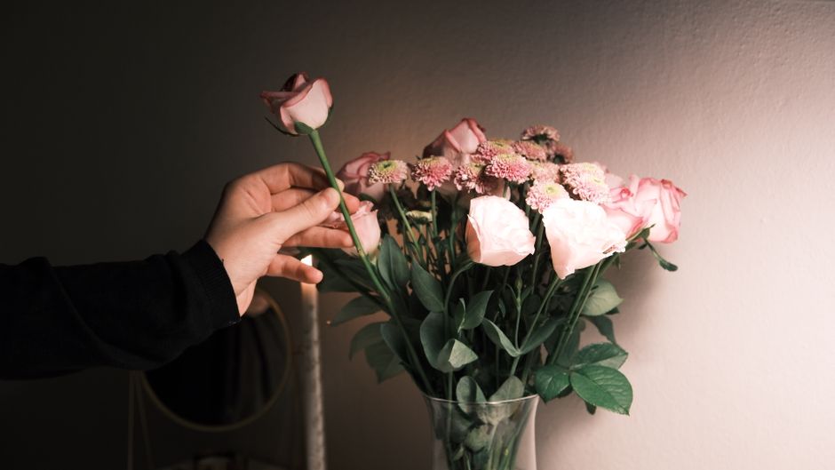A person holding a single pale pink rose above a tall clear glass vase filled with a mixed bouquet of fresh flowers, including pink roses, light pink chrysanthemums, and white blossoms. The arrangemen