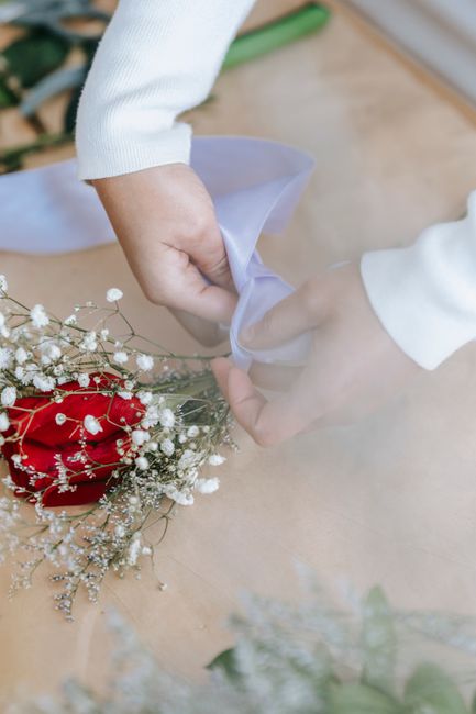 A close-up of a person's hands wrapping a bouquet of wedding flowers, featuring vibrant red roses and delicate white baby's breath, with soft, clear cellophane and a light purple ribbon. The bouquet i