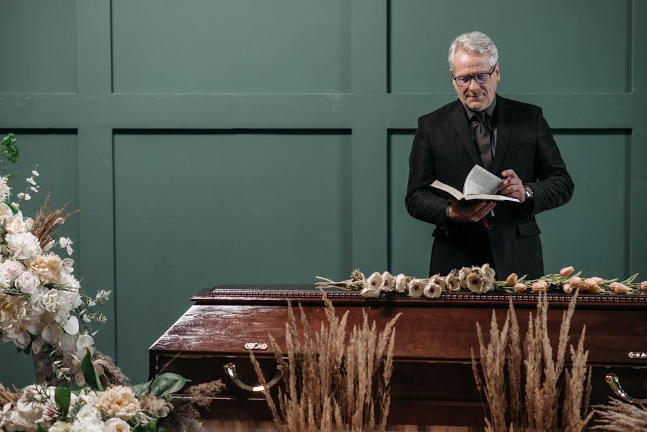 A man with grey hair and glasses, wearing a black suit and black tie, stands behind a wooden casket at a funeral service. He is holding an open book in his hands, reading aloud, with a solemn expressi