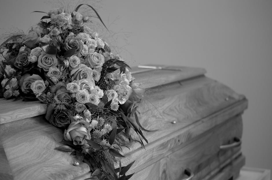 A large funeral tribute composed of a variety of fresh flowers, predominantly roses, arranged in a cascading bouquet with roses in shades of light and dark, and accented by greenery, resting on the li