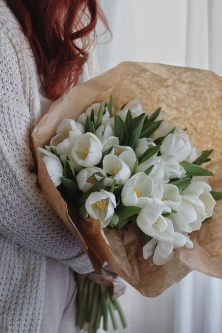 A person with long, curly red hair, wearing a light gray knitted sweater, holds a large bouquet of fresh white tulips with green stems and leaves. The tulips have open, cup-shaped petals revealing bri
