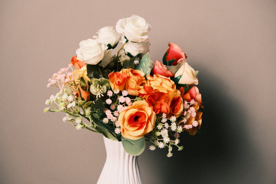 A floral arrangement featuring a bouquet of fresh roses in white, peach, and pink shades, nestled among small white and pink filler flowers and lush green foliage. The bouquet is placed in a white cer