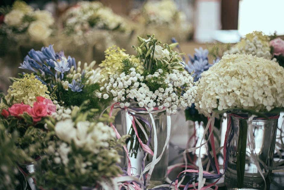 A collection of floral arrangements featuring white hydrangeas, pale pink roses, blue and white irises, and delicate white baby's breath, all arranged in transparent glass vases decorated with pink, w