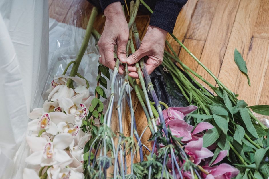 A close-up of a florist's hands arranging a variety of fresh flowers for a floral gift. The bouquet includes white orchids with pink and yellow accents, pink lilies, and green foliage, all laid out on